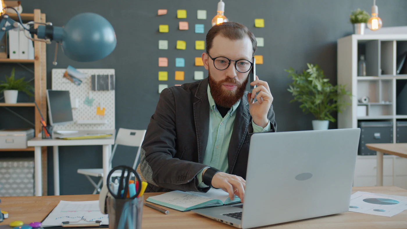 A man talking on a mobile phone while working on a laptop in an office