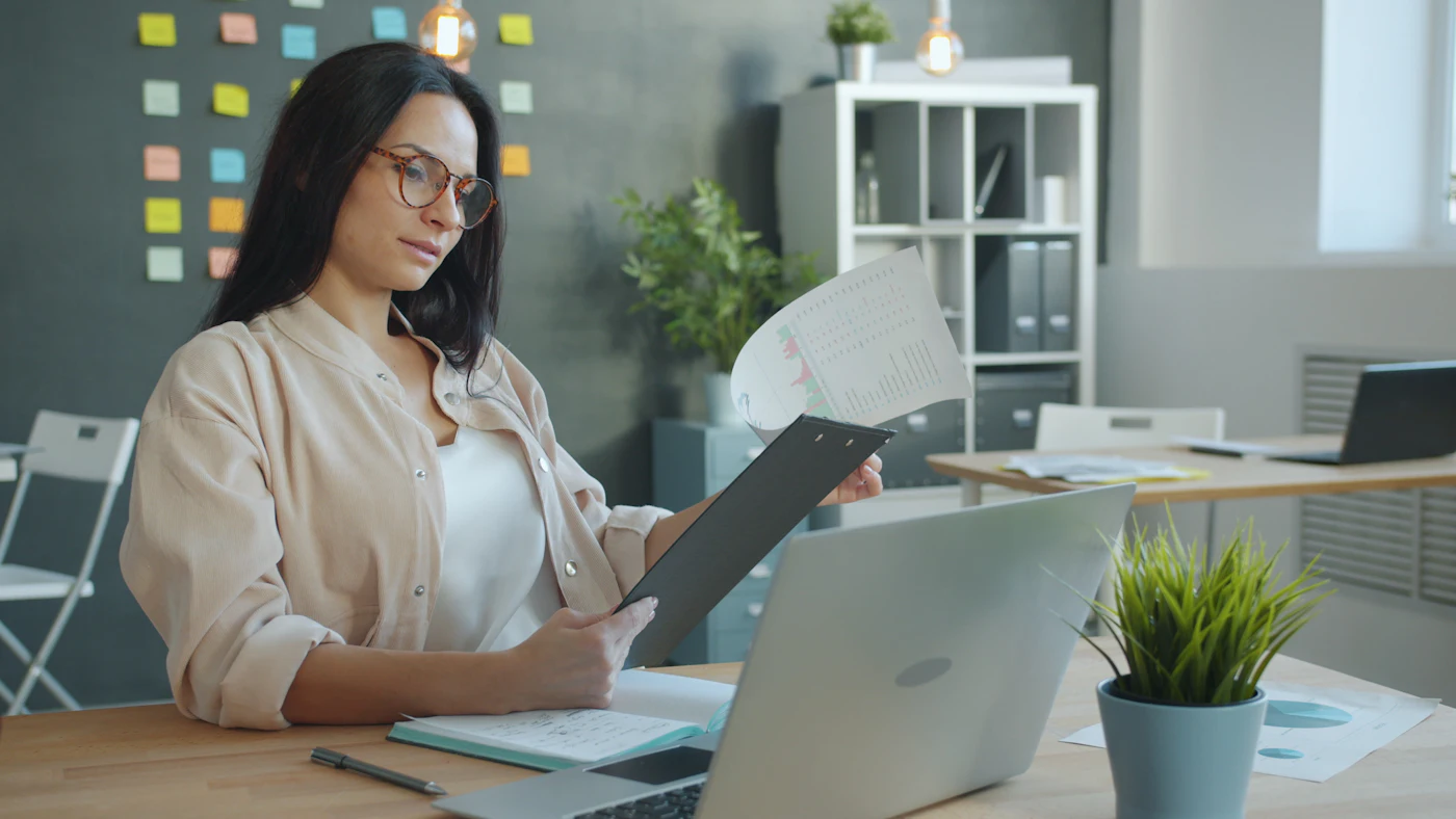 A woman looking through documents beside a laptop at an office desk