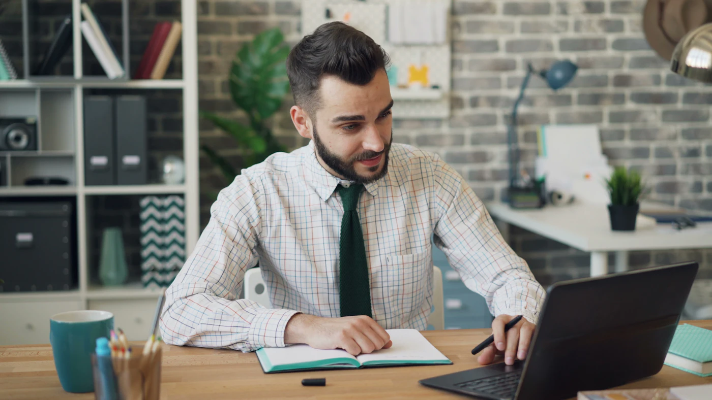 A man at an office table typing on a laptop and writing in a notebook